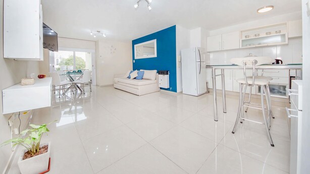 A modern kitchen with white cabinets, a blue accent wall, and a dining area with a white sofa and chairs. - Rent Corro X32 (Córdoba)