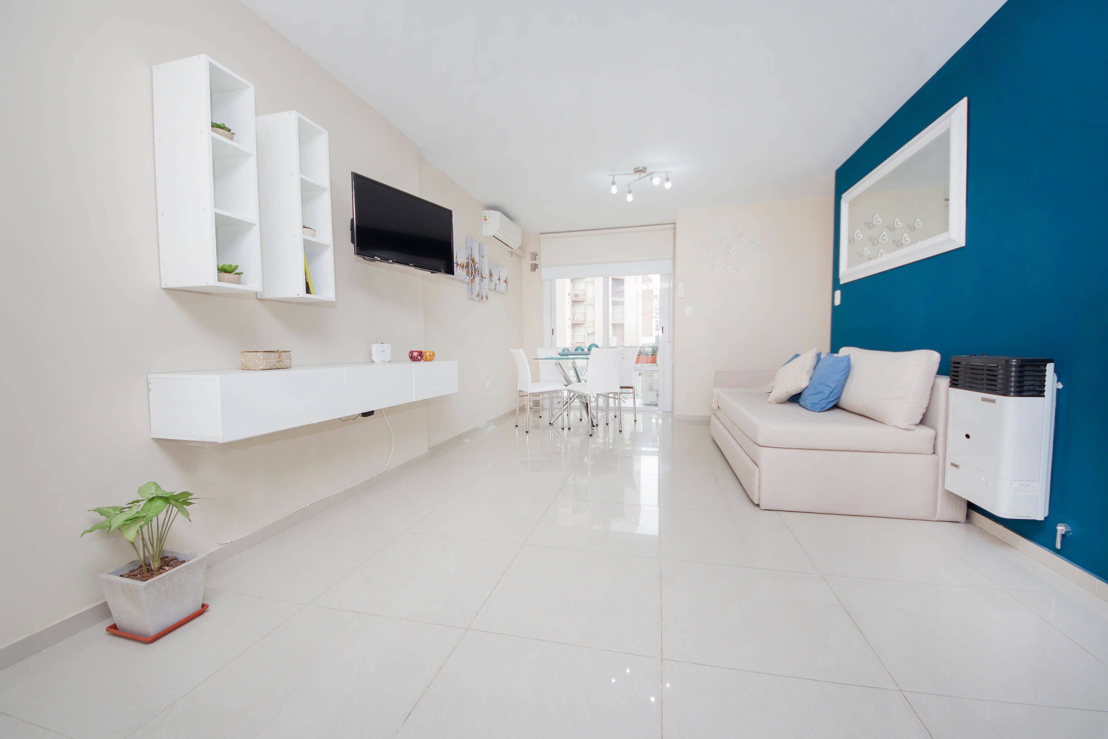 Una sala de estar moderna con un sofá blanco, un televisor montado en la pared y un comedor al fondo.