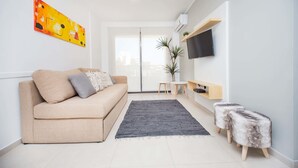 Un salon moderne avec un canapé beige, un téléviseur à écran plat, une petite table et une plante en pot.