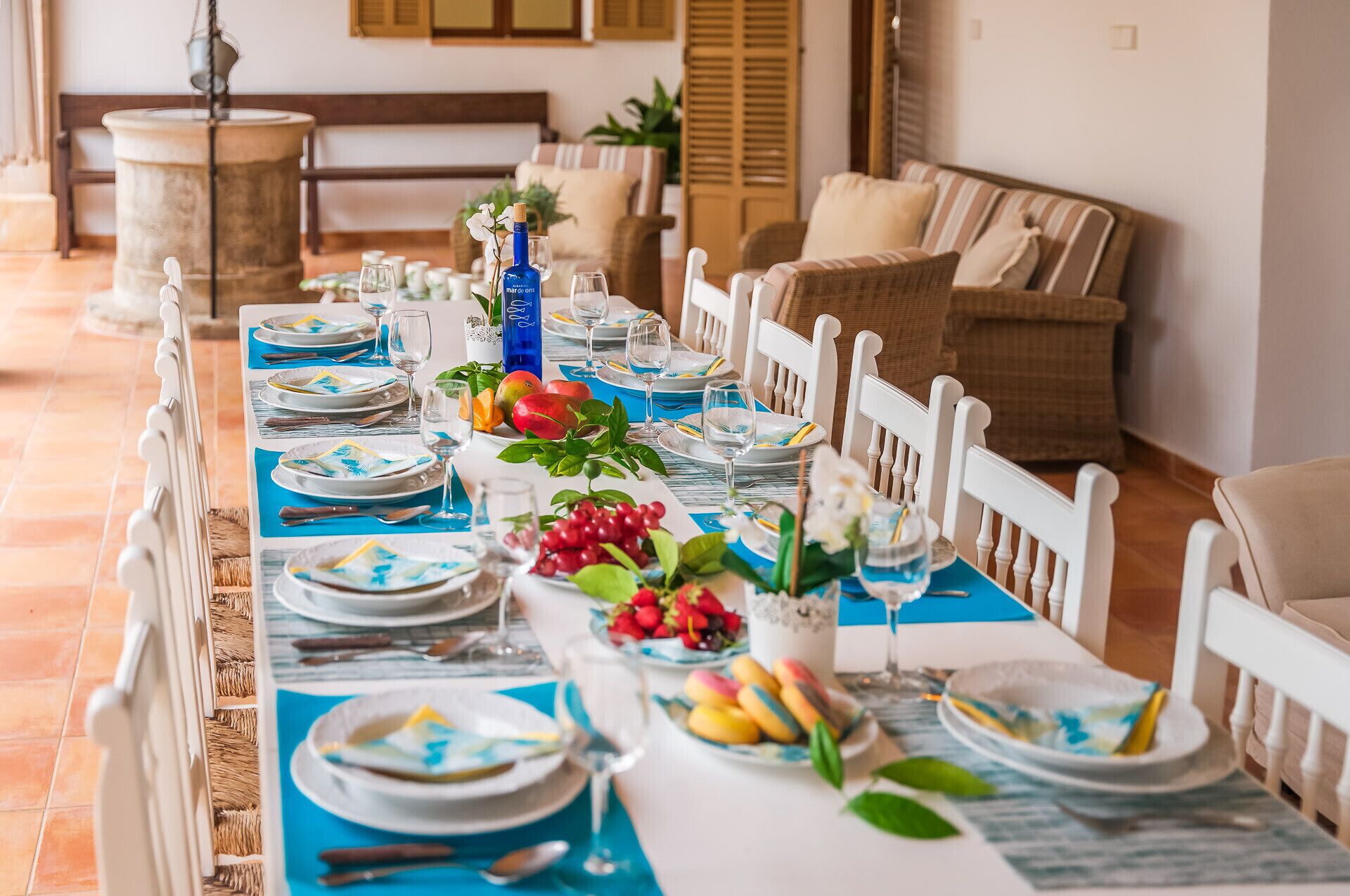 Mesa de comedor dispuesta con platos, vasos y una botella, rodeada de sillas de mimbre y un sofá en una habitación luminosa con suelo de baldosas.