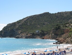 A beach with clear blue water, a rocky hillside, and several beach umbrellas.