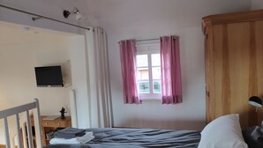 Ein Schlafzimmer mit Bett, einem Holzschrank, einem Schreibtisch mit Lampe, einem an der Wand befestigten Fernseher und einem Fenster mit Vorhängen.
