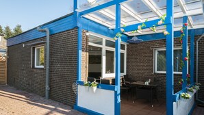 Et murstenshus med blå facade, en overdækket terrasse med et bord og stole, samt et vindue med hvid ramme.
