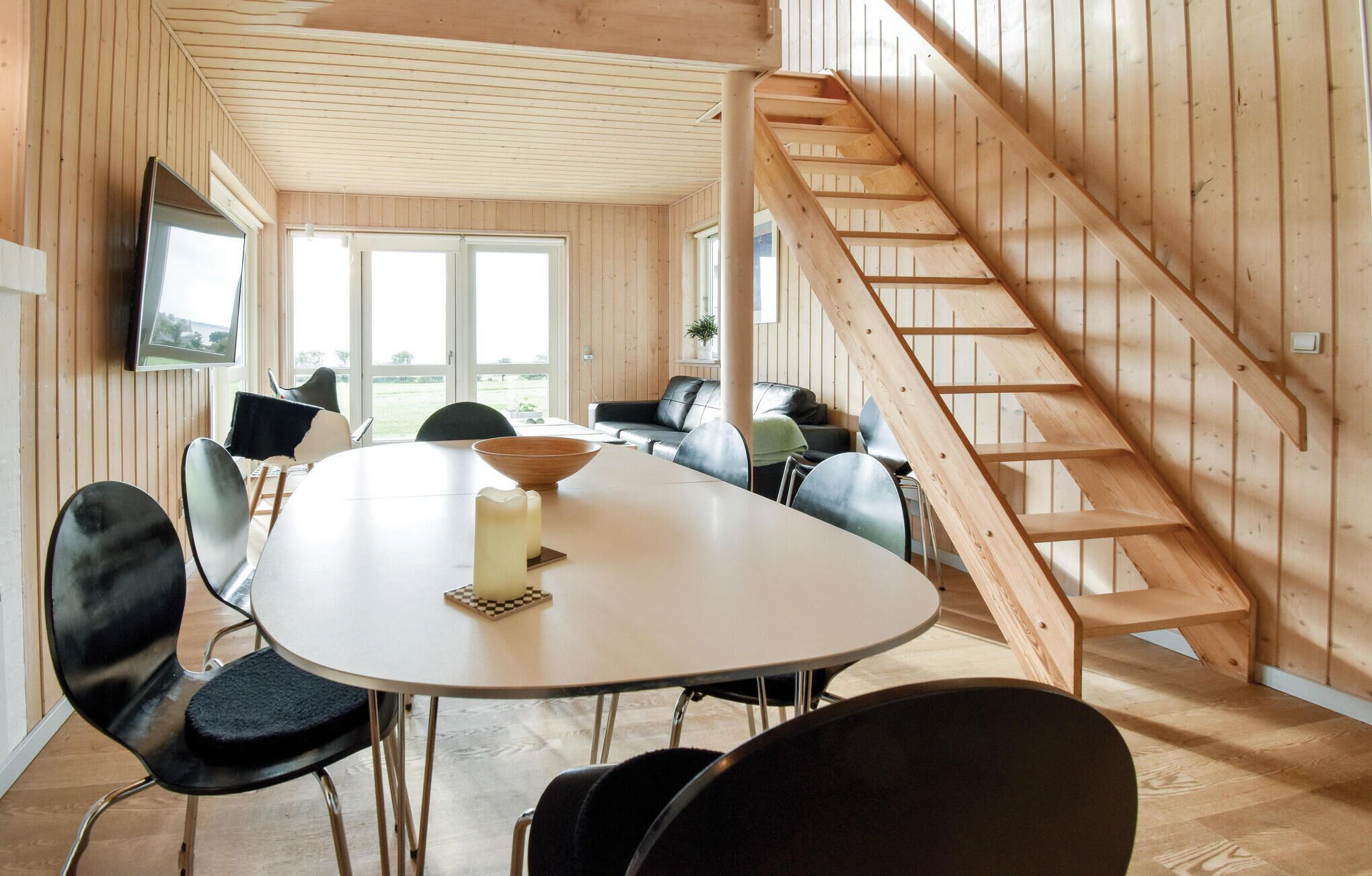 A modern interior with a wooden staircase, a round dining table with chairs, and a view of the outdoors through large windows.