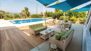 A poolside area with wooden decking, a pool, a covered seating area with a table and chairs, and a decorative vase.