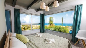 A bedroom with a large window, a bed with a green patterned bedspread, a white pillow, and a green pillow. There are two white lampshades hanging from the ceiling.