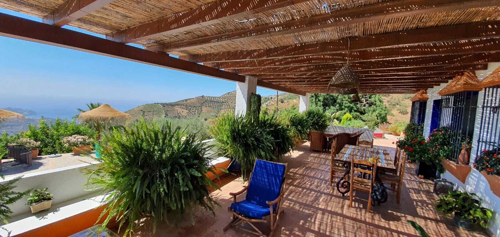 Uma  área de estar coberta ao ar livre com mesa e cadeiras, rodeada por plantas em vasos e com vista para uma paisagem deslumbrante.
