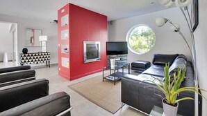 Ett modernt vardagsrum med en röd accentvägg, en svart lädersoffa, ett soffbord i glas, en platt-TV och ett stort fönster med utsikt över grönområden.