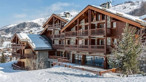 A multi-story wooden chalet with balconies, set against a snowy mountain backdrop. - Apartment for 6 with pool, spa, parking - Ski-in/Ski-out (Saint-Martin-de-Belleville)