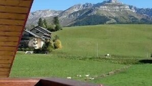A view from a wooden balcony overlooking a green field and mountains. - 6 to 7 sleeps, spacious with central fireplace, wooden top of the chalet. (Villard-De-Lans)