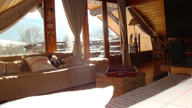 A cozy room with a wooden ceiling, a sofa with cushions, a wicker side table, and a view of a snowy landscape through large windows. - 6 to 7 sleeps, spacious with central fireplace, wooden top of the chalet. (Villard-De-Lans)