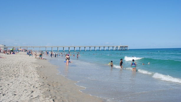 Una playa con un muelle que se adentra en el agua, gente caminando por la orilla y el océano al fondo.