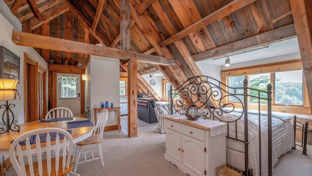 Une chambre à coucher rustique avec un plafond en bois, un lit doté d’une tête de lit en métal, une table à manger avec des chaises, et une vue sur l’extérieur grâce à de grandes fenêtres.