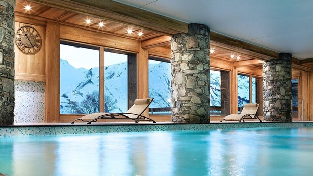 Une piscine intérieure dotée de piliers en pierre, d’un plafond en bois et de grandes fenêtres offrant une vue sur les montagnes.