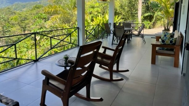 A covered outdoor seating area with wooden rocking chairs and a table, overlooking a lush green landscape. - Villas Lambeau #1 Ocean View  - Fiber Optic / 1BR 1 BA (Uvita)
