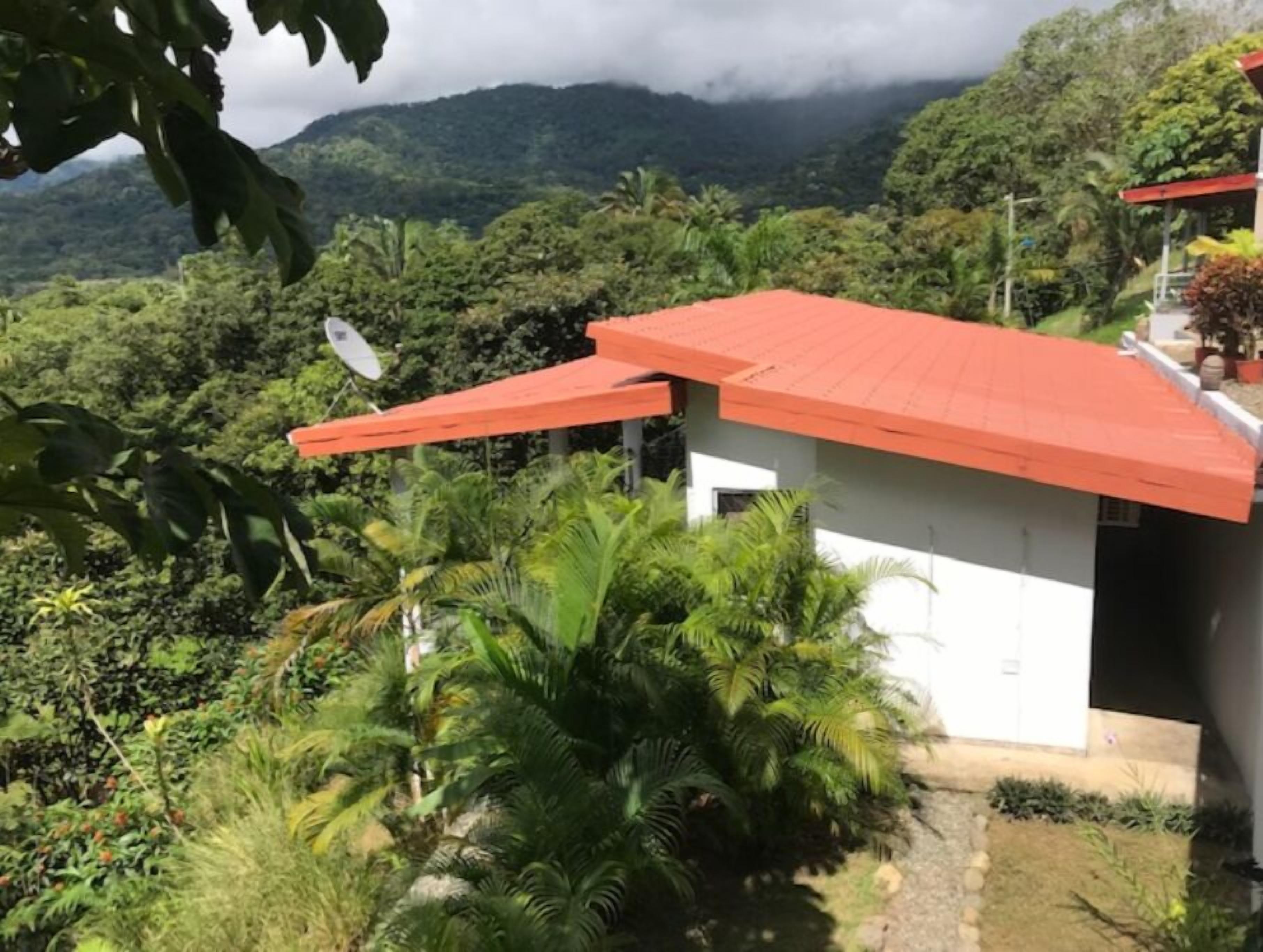 Un pequeño edificio de techo rojo, rodeado de abundante vegetación y montañas al fondo.