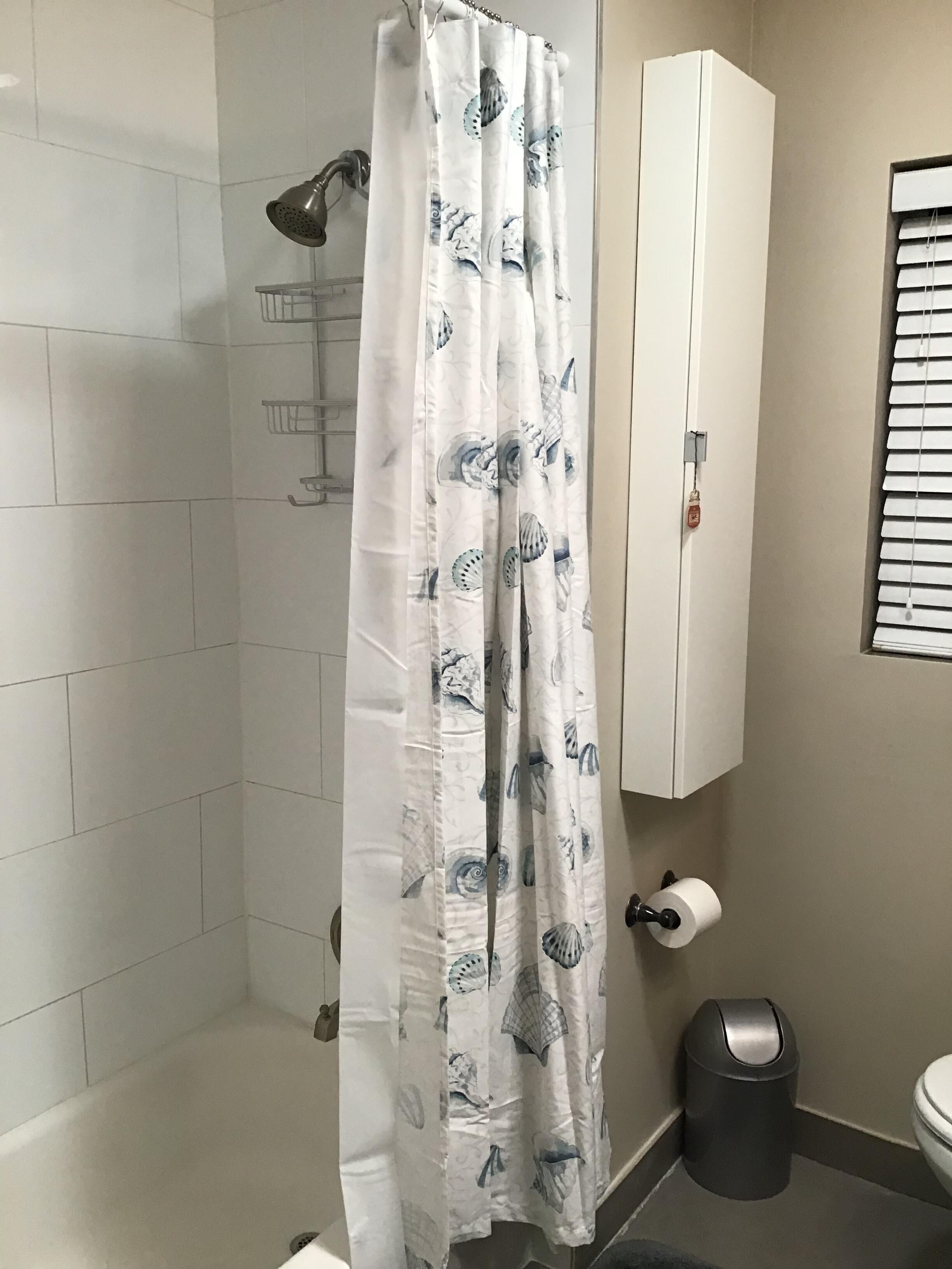 A bathroom with a shower curtain featuring a shell pattern, a toilet, a trash can, and a towel rack.