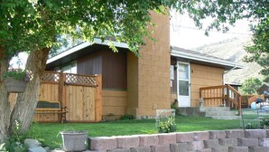 A two-story house with a brick exterior, a wooden deck, and a front yard with a stone wall and a tree.