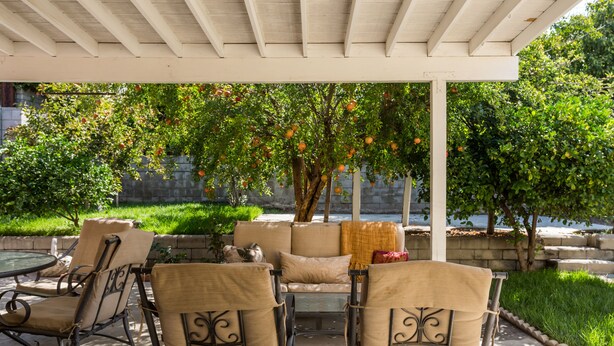 En udendørs patio med overdækket siddeområde, herunder en sofa og stole, under en pergola med frugtbærende træer.