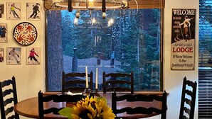 Ein Essbereich mit Tisch, Stühlen, einer Chandelüster, einem Fenster mit Blick auf Bäume und einem Willkommensschild für die Apres-Ski Lodge.