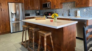 Eine moderne Küche mit Holzausstattung, Edelstahlgeräten, einer großen Kochinsel mit Barhocker und einer Spritzschutzwand aus strukturierten Fliesen.
