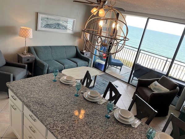 Espaço de estar moderno com ilha de cozinha, mesa de jantar e assentos. Uma janela grande oferece vista para o oceano.