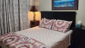 Una cama bien hecha con ropa de cama estampada, una lámpara de noche, un cuadro enmarcado en la pared y una mesita de noche con una caja de pañuelos.