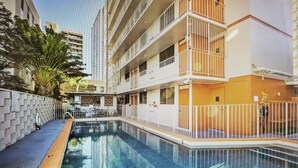 Una piscina rodeada por una valla blanca, con un edificio de varios pisos al fondo.