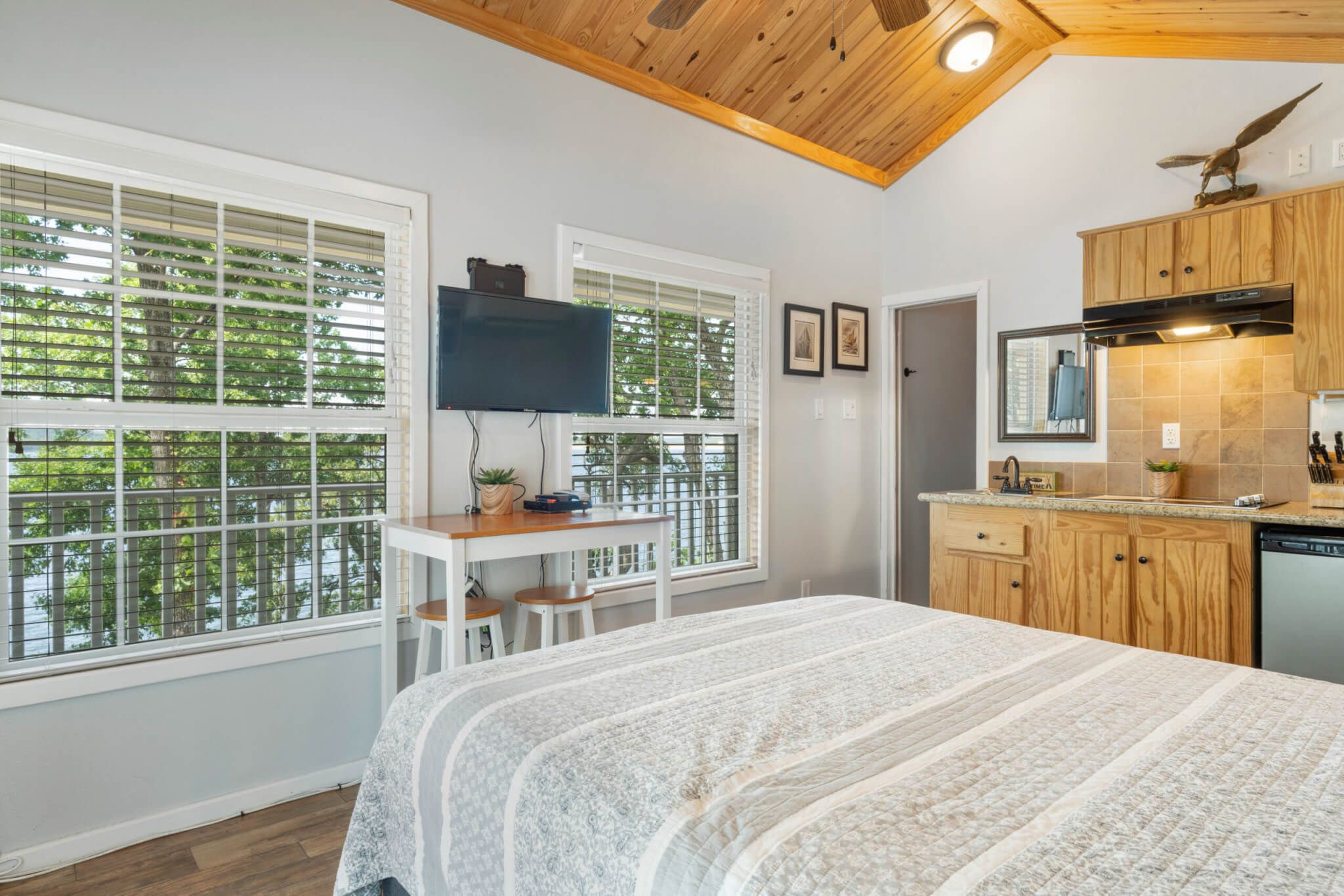 A compact room with a bed, a desk, a TV, and a kitchen area with wooden cabinets and a dishwasher.