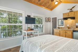 Habitación funcional con cama, escritorio, televisor y una cocina americana con muebles de madera y lavavajillas.