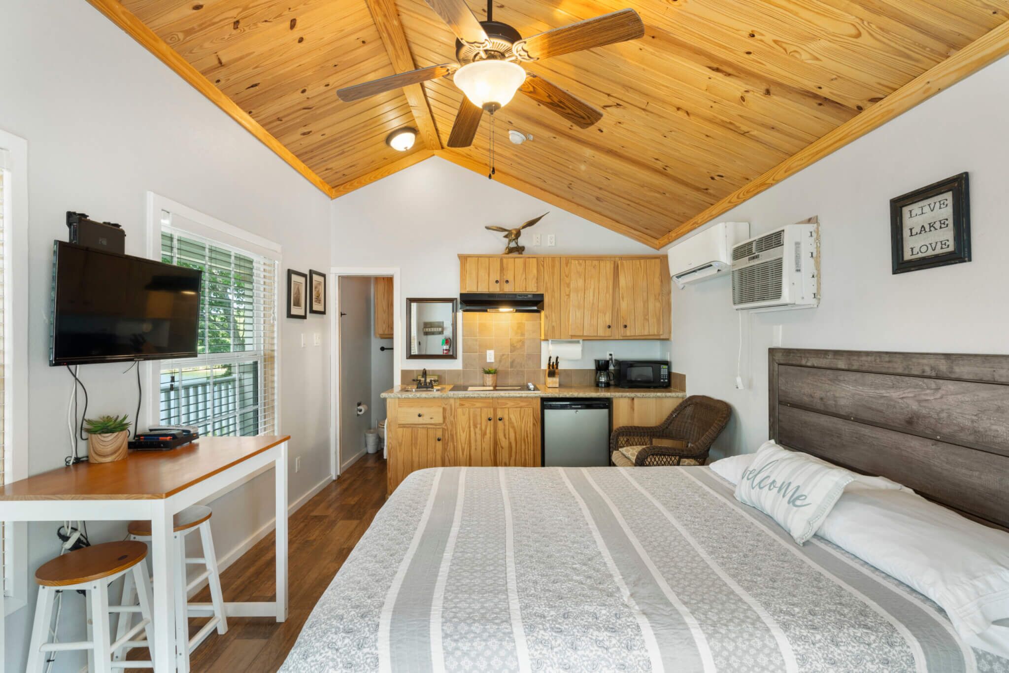 A bedroom with a large bed, a wooden ceiling, a desk with a TV, a kitchen area, and a window with blinds.