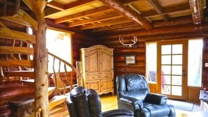 A rustic cabin interior with wooden walls, a leather armchair, a wooden cabinet, and a staircase.