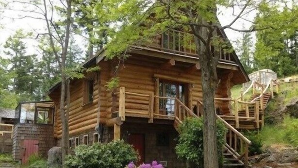 A log cabin with a wooden staircase leading to a deck, surrounded by greenery and a stone wall.