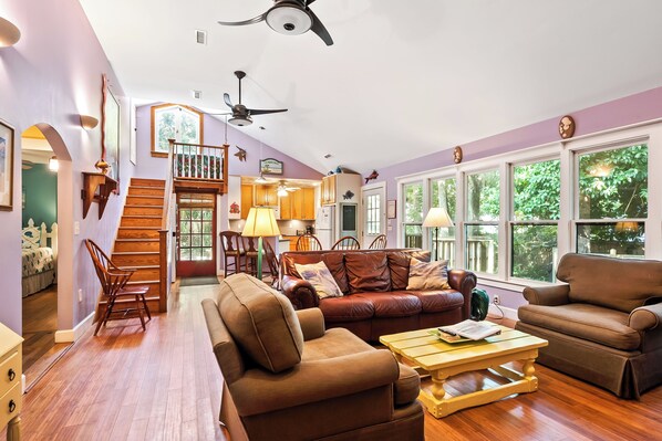 A spacious living room with a staircase, leather sofas, wooden flooring, and large windows.