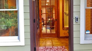 A wooden door leading into a room with a patterned rug, a window with a view of greenery, and a doorbell.