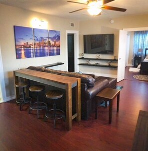 A modern living room with a wooden bar and stools, a leather sofa, and a flat-screen TV mounted on the wall. - Modern 2/1 apt near Midtown Miami and South Beach (Miami)