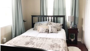 Ein Schlafzimmer mit Bett, Vorhängen, einem Nachttisch mit Lampe und einer kleinen Pflanze.