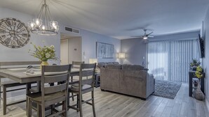 Een moderne woonkamer met een grijze bank, een eettafel met stoelen, een grote wandklok en een plafondventilator.