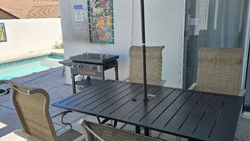 An outdoor patio with a table and chairs, a grill, and a pool in the background.
