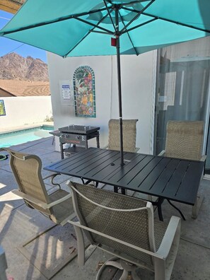 An outdoor patio with a table and chairs, a grill, and a pool in the background.