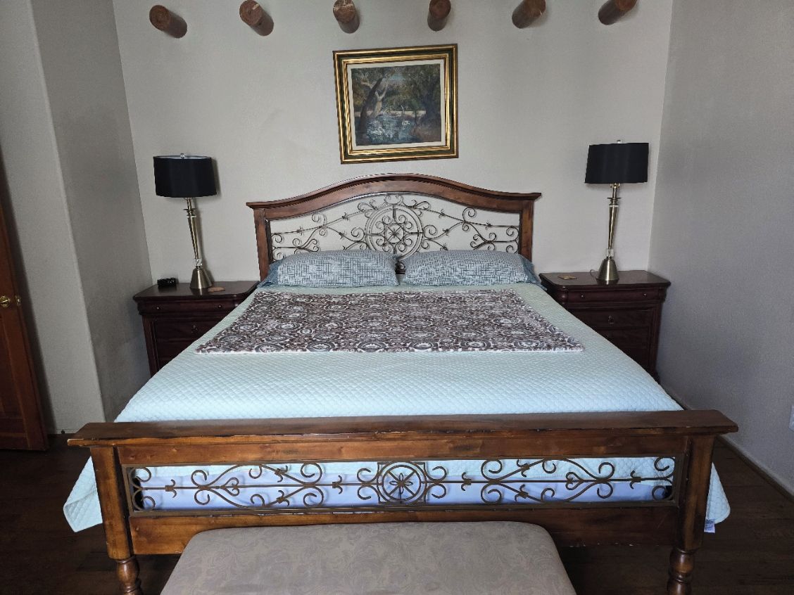 Een slaapkamer met een houten bed, twee nachtkastjes met lampen, een lijst met een afbeelding aan de muur en een bankje aan het voeteneinde van het bed.