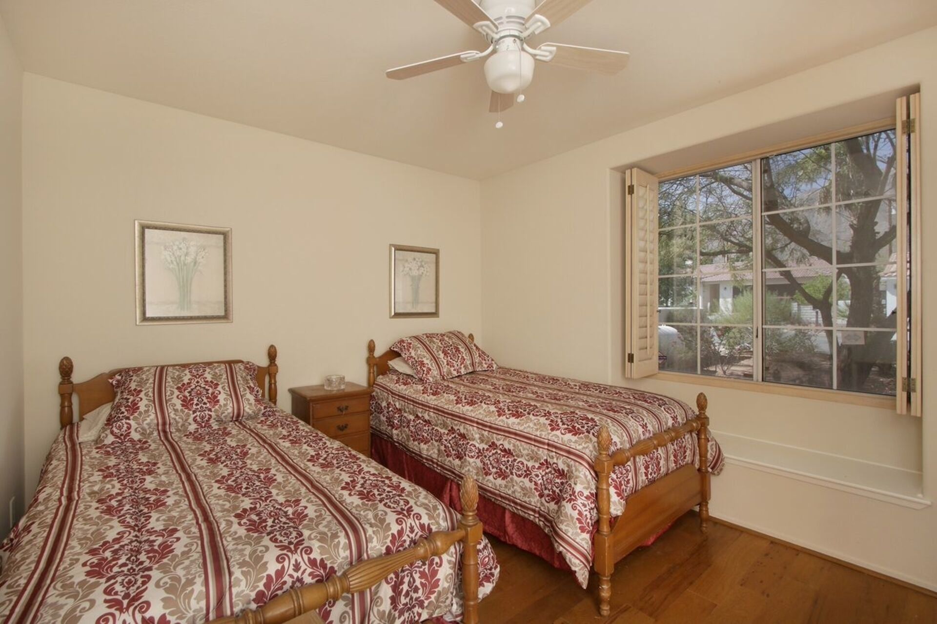 A bedroom with two wooden beds, a ceiling fan, and a large window with a view of trees and a house.