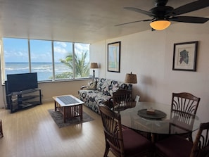 A living room with a view of the ocean, a television, a sofa, a coffee table, and a dining table with chairs.