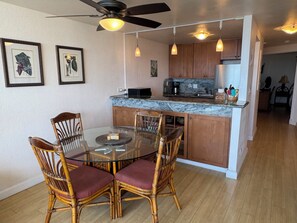 A dining area with a glass-top table and four chairs, a kitchen with wooden cabinets and a marble countertop, and a ceiling fan.