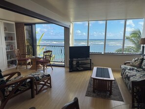 A living room with a large window overlooking the ocean, a television, and various seating arrangements.