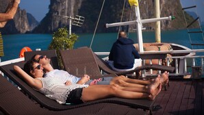 A couple relaxing on a sun lounger with a view of a mountainous coastline. - Golden Star Cruise (Tuan Chau)