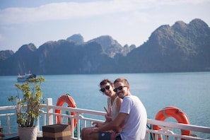 A couple sitting on a boat deck with a view of a mountainous coastline and a ship in the distance.