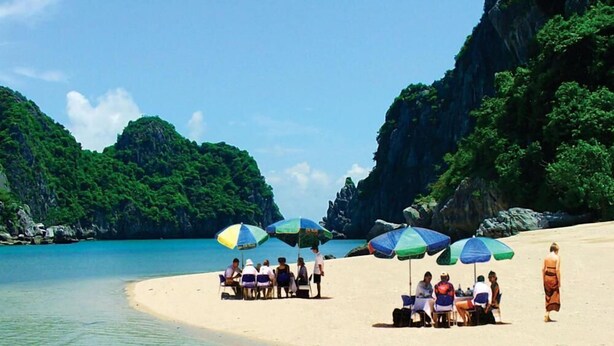 A beach with people under umbrellas, a clear sky, and lush greenery in the background. - Golden Star Cruise (Tuan Chau)