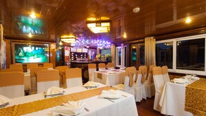 A restaurant with multiple tables set for dining, wooden ceiling, and a bar area in the background. - Golden Star Cruise (Tuan Chau)