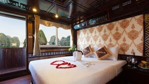 A hotel room with a large bed, a window with curtains, a nightstand with a fruit bowl, and a wall clock. - Golden Star Cruise (Tuan Chau)
